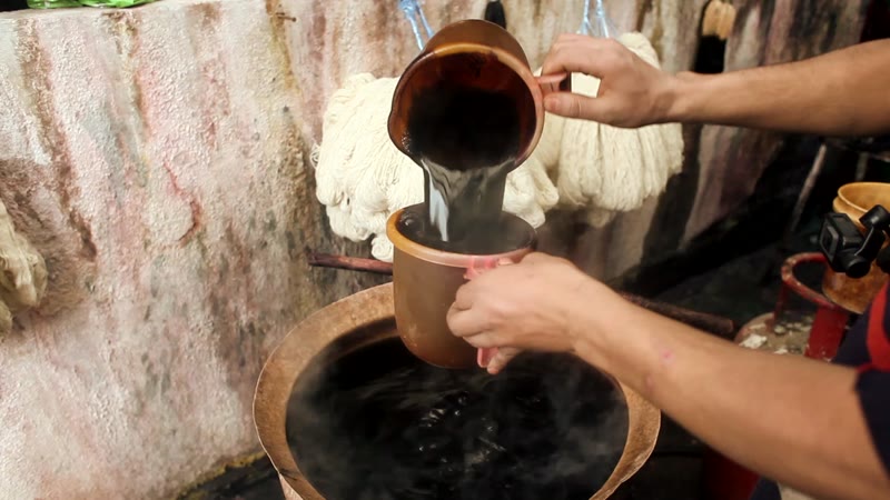 Dye poured from one copper pot into another beside a dyeing vat, with pale skeins of yarn hanging behind